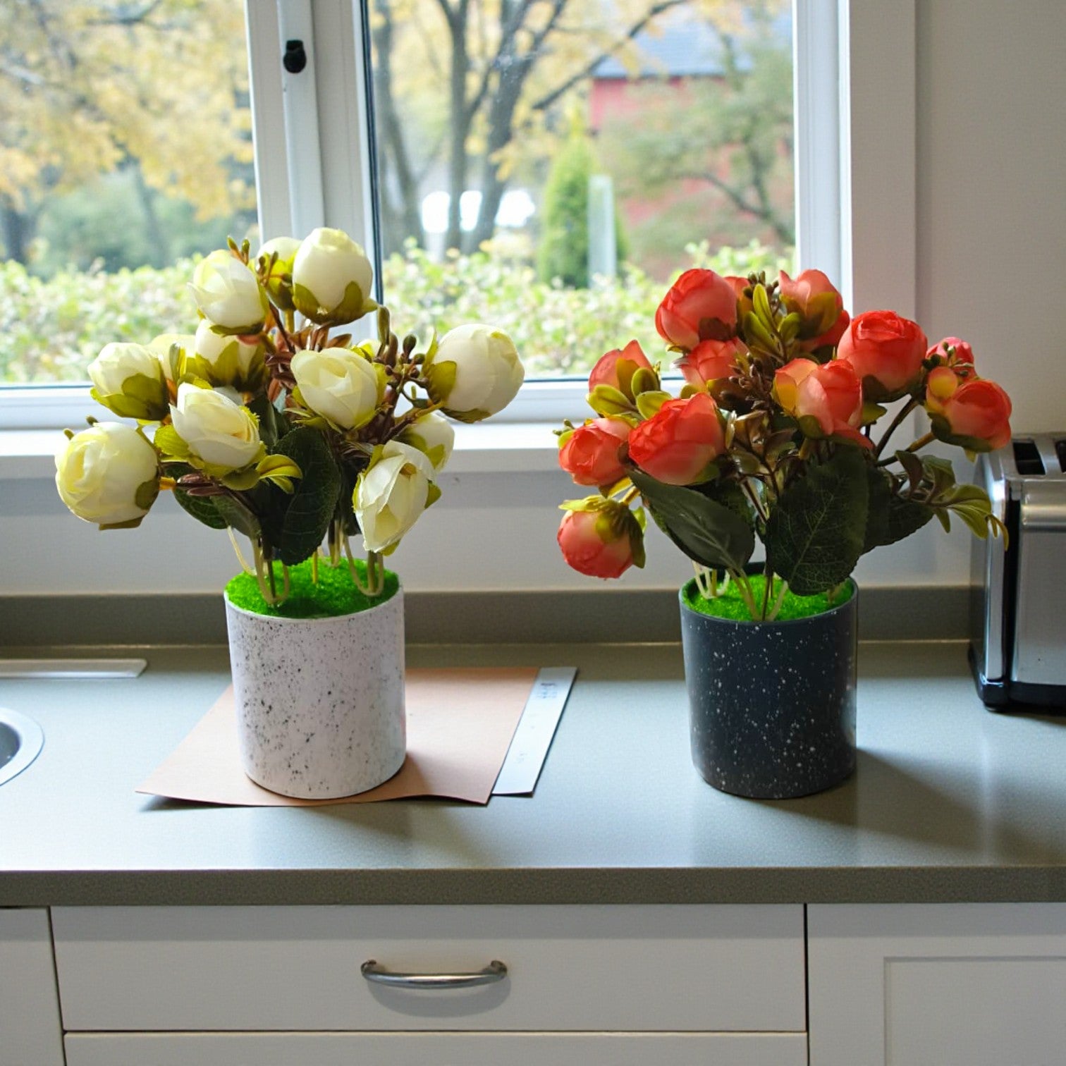 Set of two small artificial rose bud plants with white flowers in a light grey pot and orange flowers in a dark charcoal pot.