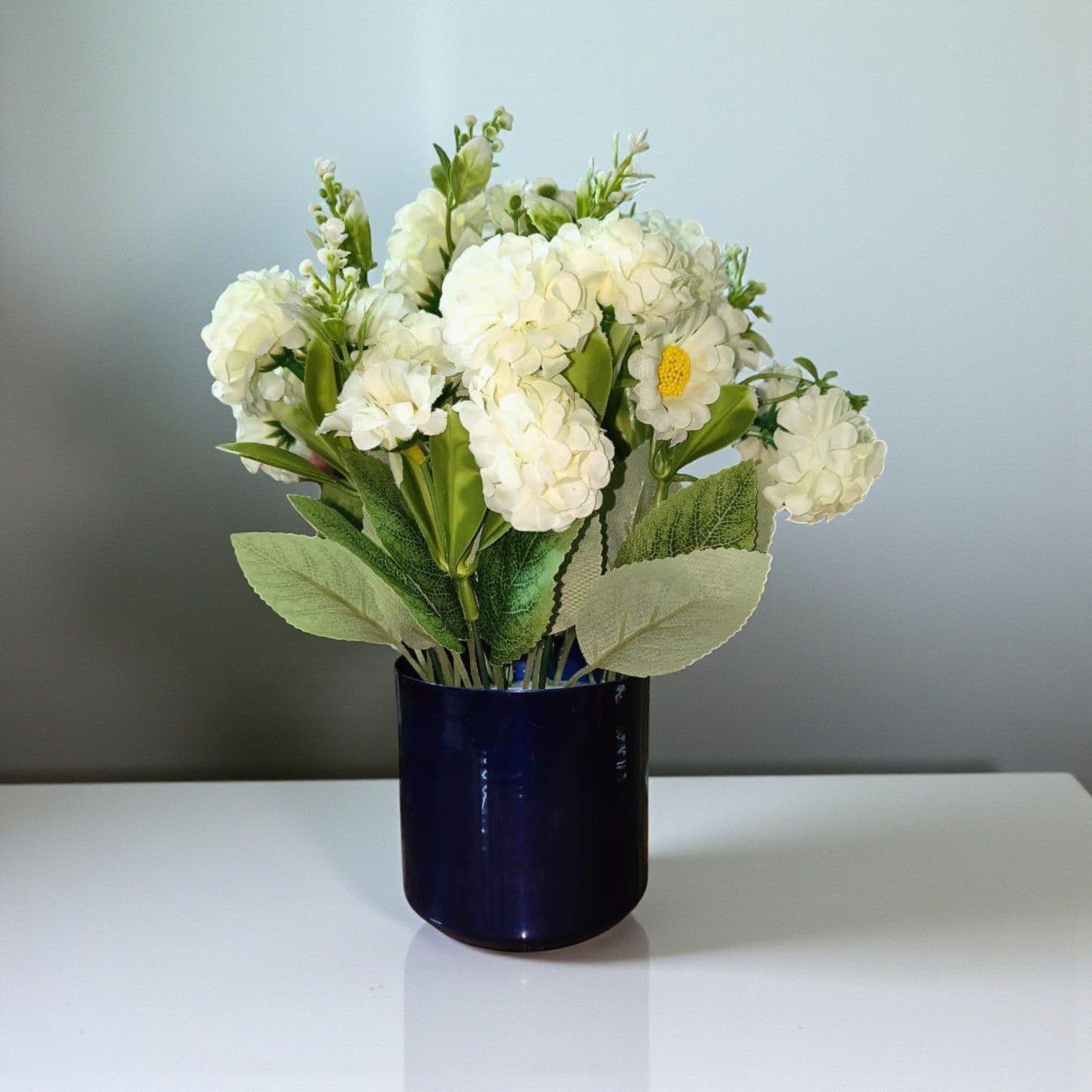 Artificial white hydrangea flowers in a cylindrical deep blue metal pot.